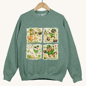 May include: Green sweatshirt featuring four square illustrations of cats celebrating St. Patrick's Day. Each cat wears a green hat and is surrounded by shamrocks, rainbows, and pots of gold. The sweatshirt is hanging on a wooden hanger.