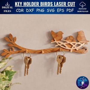 Może przedstawiać: Drewniany uchwyt na klucze z motywem gałęzi, ptakami wyciętymi laserowo i gniazdem. Uchwyt na klucze ma trzy haczyki i zawiera tekst "KEY HOLDER BIRDS LASER CUT". Klucze są przymocowane do złotych kółek.