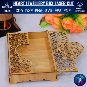 May include: A laser-cut wooden heart-shaped jewelry box with intricate geometric and floral designs. The box is open, revealing the interior, and features the text "HEART JEWELLERY BOX LASER CUT" and file type options. The box is made of light-colored wood.
