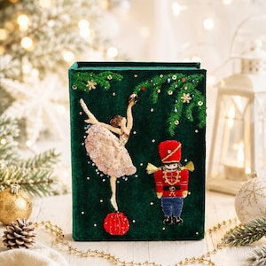 May include: A dark green velvet book cover featuring intricate embroidery of a ballerina in a beige tutu and a red nutcracker soldier. The design includes green pine branches, small gold stars, and scattered beads on a deep green fabric surface.