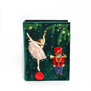 May include: A green velvet box with a ballerina and a nutcracker embroidered on it. The ballerina is wearing a pink tutu and holding a red ornament. The nutcracker is wearing a red and blue uniform. The box is decorated with green pine branches and gold glitter.