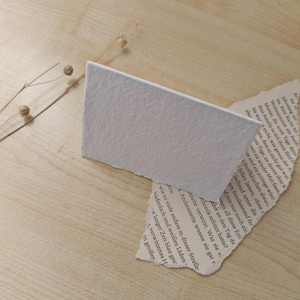 Könnte beinhalten: Eine rechteckige weiße Karte mit strukturierter Oberfläche, schräg platziert auf einem Stück zerrissenem Papier mit gedrucktem deutschem Text. Ein dünner, getrockneter Zweig mit kleinen runden Knospen erstreckt sich über die obere linke Ecke.