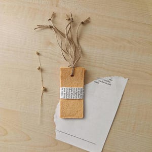 Könnte beinhalten: Ein handgefertigter, rechteckiger Lesezeichen mit natürlicher, strukturierter Oberfläche. Es zeigt einen Streifen bedrucktes Papier und ist mit Bindfaden gebunden. Ein Zweig getrockneter Blumen und zerrissenes Papier sind ebenfalls vorhanden.