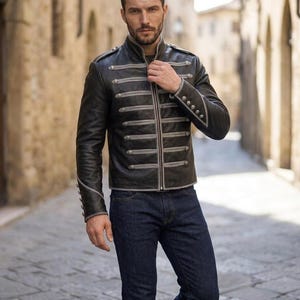 Könnte beinhalten: Eine schwarze Lederjacke mit hohem Kragen, silberfarbenen geflochtenen Verzierungen im Militärstil und dekorativen Metallknöpfen an der Vorderseite und an den Ärmelbündchen. Die Jacke wird mit einer dunkelblauen Jeans kombiniert.