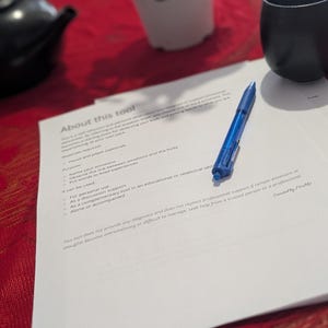 Peut inclure: Un stylo bleu est posé sur une feuille de papier blanche intitulée "About this tool". Le papier contient des puces et du texte. Une théière noire, une petite plante en pot et une tasse noire sont en arrière-plan sur une nappe rouge.