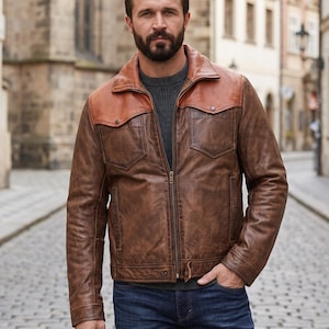 May include: A brown leather jacket featuring a contrasting tan leather yoke across the shoulders, a full-length front zipper, two chest pockets with buttoned flaps, and a ribbed dark gray sweater worn underneath with dark blue denim jeans.