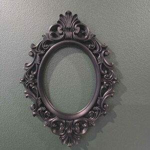 May include: A black oval picture frame with elaborate scrollwork and a dark, matte finish. The frame is empty in the centre. It is set against a textured, muted green background.