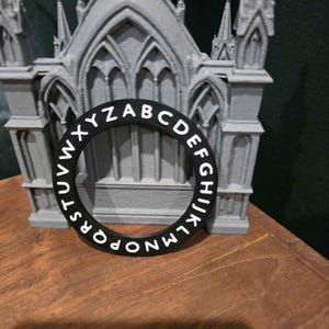 Peut inclure: Un objet circulaire noir avec des lettres blanches formant l'alphabet, placé devant une maquette architecturale grise. Les lettres sont espacées uniformément autour du cercle. La maquette est une structure grise détaillée.
