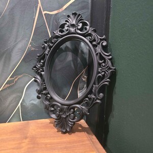 May include: A black, oval mirror with an elaborate, baroque-style frame. The frame has detailed scrollwork and floral patterns. The mirror is placed on a wooden surface, with a dark green and gold abstract background.