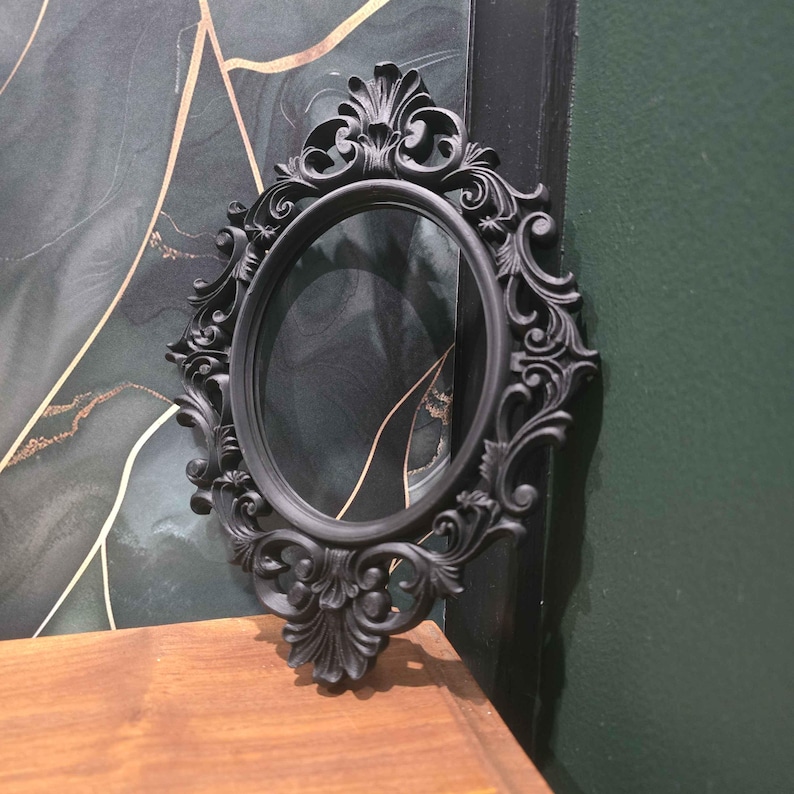 May include: A black, oval mirror with an elaborate, baroque-style frame. The frame has detailed scrollwork and floral patterns. The mirror is placed on a wooden surface, with a dark green and gold abstract background.