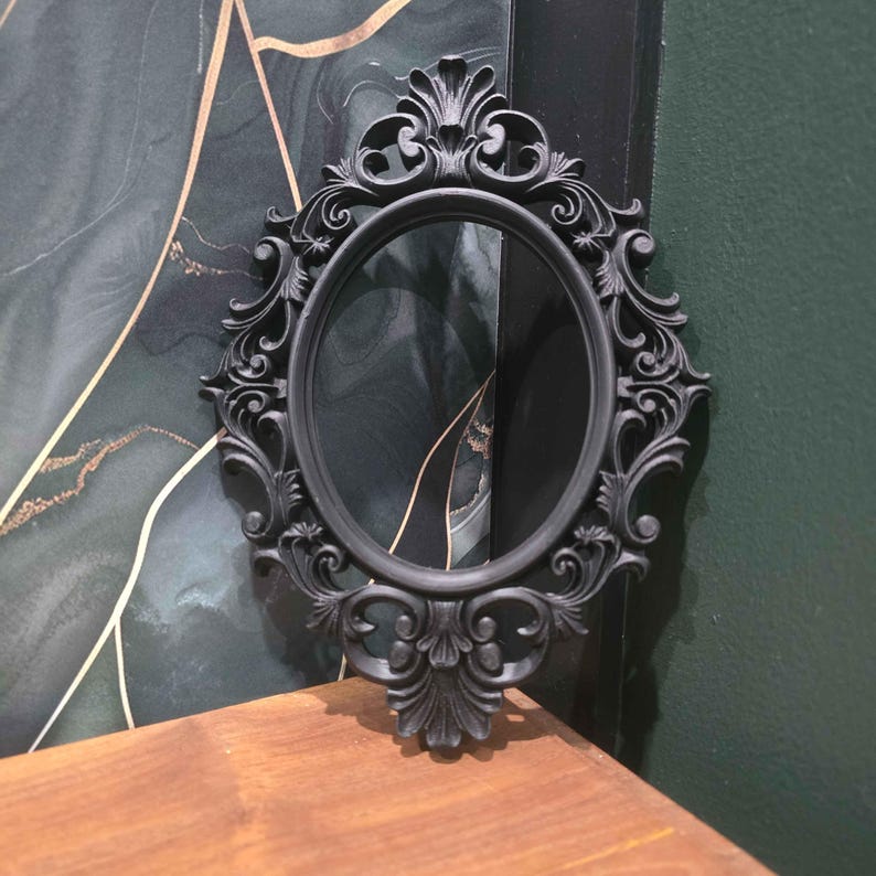May include: Black oval mirror with elaborate, swirling frame detailing. The matte black frame has a decorative top and bottom design. The mirror is set against a dark green and gold abstract background.