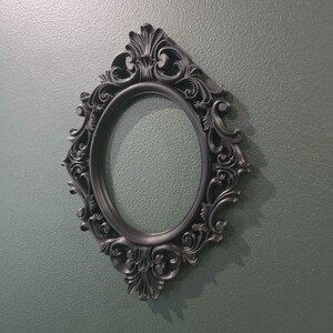May include: An oval, black picture frame with elaborate, swirling decorative details. The frame is mounted on a textured, muted green wall, creating a shadow. The frame is a decorative item.