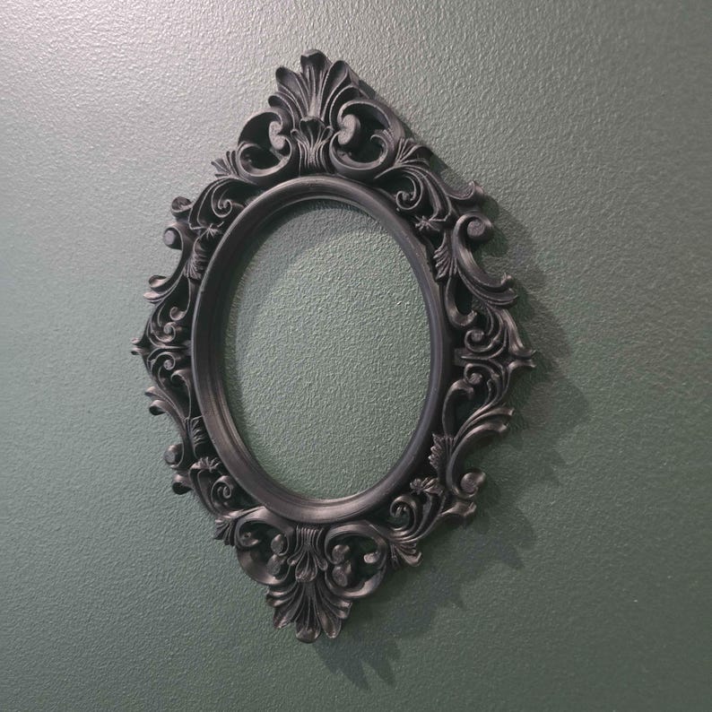 May include: An oval, black picture frame with elaborate, swirling decorative details. The frame is mounted on a textured, muted green wall, creating a shadow. The frame is a decorative item.