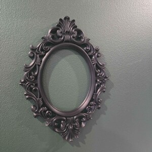 May include: An oval black picture frame with elaborate scroll detailing. The frame has a dark, matte finish and is empty. A decorative item for displaying photographs or artwork. Wall decor.