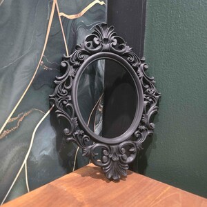 May include: A black, oval-shaped mirror with elaborate scroll detailing. The mirror is framed in black and is positioned against a backdrop of green and gold patterned wallpaper and a dark green wall. It rests on a wooden surface.