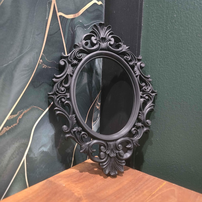 May include: A black, oval-shaped mirror with elaborate scroll detailing. The mirror is framed in black and is positioned against a backdrop of green and gold patterned wallpaper and a dark green wall. It rests on a wooden surface.