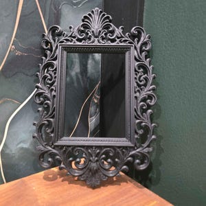 以下が含まれることがあります： 詳細な渦巻き模様と装飾的な上部と下部を備えた、黒色の長方形の装飾鏡。 鏡は、金色のアクセントが付いた濃い緑色の壁を背景に、木製の表面に表示されています。