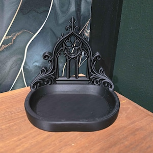 May include: Black decorative tray with an ornate gothic design. The tray features a central oval basin and an arched back with intricate tracery. The tray is made of a solid black material and sits on a wooden surface.