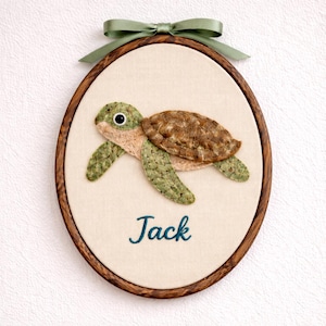 Puede incluir: Adorno de pared ovalado enmarcado con un diseño de tortuga hecho a mano. La tortuga está hecha de tela texturizada en tonos verdes, marrones y beige. El nombre "Jack" está bordado en verde azulado debajo. Un lazo de cinta verde está atado en la parte superior.