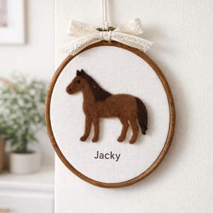 May include: A brown felt horse embroidery hoop with the name "Jacky" in black text. The horse is centered on a white fabric, framed by a wooden hoop, and topped with a cream-coloured bow.