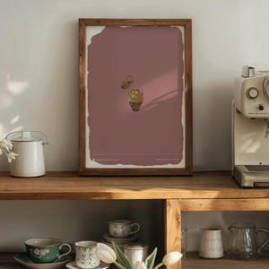 Puede incluir: Una impresión artística enmarcada con un fondo rosa polvoriento y dos ilustraciones de urnas doradas. El marco de madera rodea la impresión, que se exhibe en un estante de madera. También se ven una tetera blanca y una cafetera.