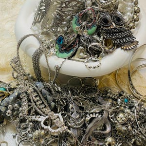 May include: A collection of silver-toned jewelry, including necklaces, bracelets, rings, and earrings, overflowing from a white bowl. The assortment features various designs, such as an owl pendant, leaf shapes, and turquoise accents. The jewelry is displayed on a cream-colored lace surface.