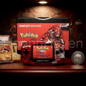 May include: A red translucent Game Boy Advance handheld console featuring a Groudon graphic, displaying the Pokémon Ruby Version title screen. Behind it sits the original game box with matching artwork, alongside a Pokémon trading card.