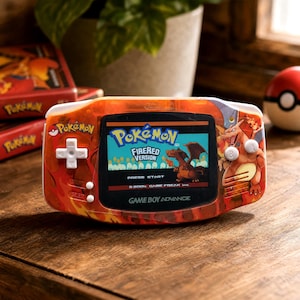Puede incluir: Una consola portátil Game Boy Advance con carcasa naranja translúcida, gráficos de llamas y una ilustración de Charizard. La pantalla muestra el título Pokémon FireRed Version con el texto Press Start y 2004 Game Freak inc.