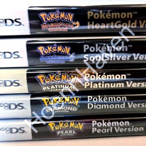 Puede incluir: Una pila de cinco cajas de juegos de Nintendo DS con las versiones Pokémon HeartGold, SoulSilver, Platinum, Diamond y Pearl. Cada caja tiene un lomo blanco con el logotipo de Nintendo DS y una sección negra con el título del juego.