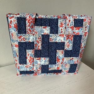 May include: A handmade tote bag with a patchwork design. The bag features a combination of navy blue fabric with a floral pattern and solid navy blue squares. The bag is sitting on a white surface.