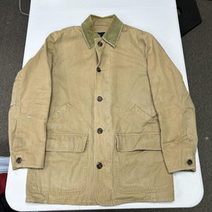 Könnte beinhalten: Eine beigefarbene Utility-Jacke mit einem kontrastierenden olivgrünen Cordkragen, einer durchgehenden Knopfleiste auf der Vorderseite und zwei großen aufgesetzten Taschen in der Taille. Die langen Ärmel haben geknöpfte Manschetten und ein robustes Design.
