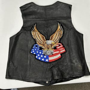 May include: A black leather vest featuring a large embroidered patch on the back. The patch depicts a brown and white eagle with spread wings perched above a motorcycle engine, set against a flowing American flag banner with stars and stripes.