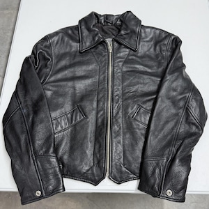 Puede incluir: Chaqueta de cuero negro con cierre de cremallera, dos bolsillos delanteros y cuello. La chaqueta tiene mangas largas con puños abotonados. El cuero parece texturizado y la chaqueta está sobre una superficie blanca.