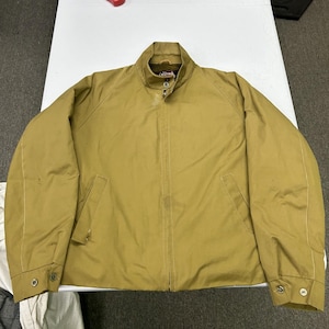 May include: A tan-coloured jacket featuring a full-length front zip, a stand-up collar, and long sleeves with buttoned cuffs. The garment includes two slanted front pockets and a visible brand label located inside the collar area.
