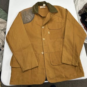 May include: A tan canvas field jacket featuring a dark olive green corduroy collar, a quilted shoulder patch on the right side, three front buttons, a single chest pocket, and two large lower pockets. The jacket has long sleeves and a structured fit.