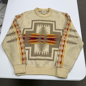 May include: A cream-colored long-sleeve sweater featuring a large, centered geometric cross pattern in shades of brown, orange, and yellow. The sleeves are decorated with vertical bands of matching geometric motifs, and the garment has a ribbed crew neckline.