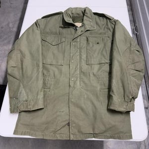 May include: An olive green military-style jacket with a button-up front, two chest pockets, and two lower pockets. The jacket has a high collar and long sleeves with buttoned cuffs. The fabric appears worn, suggesting a vintage or used condition.