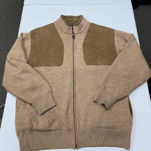 May include: A tan knit zip-up sweater featuring a mock neck collar and brown quilted shoulder patches with a geometric stitched pattern. The garment includes a full-length front zipper, ribbed cuffs, and a ribbed hem, providing a structured look.