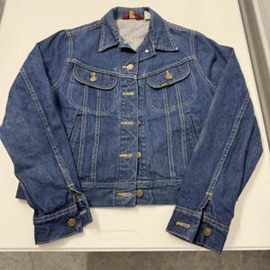 May include: A vintage denim jacket in a medium blue wash. The jacket has a classic design with button fastenings, two chest pockets, and buttoned cuffs. The jacket features gold-coloured buttons and stitching.