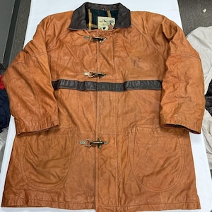 Può includere: Una giacca in pelle color cuoio con colletto marrone scuro, una striscia orizzontale in pelle marrone scuro sul petto e tre chiusure a levetta in metallo. L'interno è foderato con un tessuto a quadri verde, rosso e giallo con una piccola etichetta.
