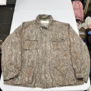 May include: A long-sleeve button-up jacket featuring a brown and tan tree bark camouflage pattern. The jacket includes a standard collar, a front button closure, and four large rectangular pockets on the front, each with a buttoned flap closure.