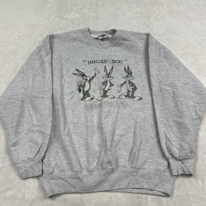 May include: A heather gray crewneck sweatshirt featuring a graphic print of three Bugs Bunny characters in different poses. Above the characters, the text reads "BUGSY BOY" in a black serif font, with additional small text printed below the figures.