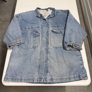 May include: Light blue denim jacket with two chest pockets and two lower pockets. The jacket has a button-up front, a collar, and cuffed sleeves. The fabric shows signs of wear and tear, giving it a vintage look. The jacket is laid flat on a white surface.