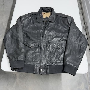 Könnte beinhalten: Schwarze Lederjacke mit Kragen, zwei Vordertaschen und geripptem Saum und Bündchen. Die Jacke hat einen Knopfverschluss und ein helles Futter, das am Kragen sichtbar ist.