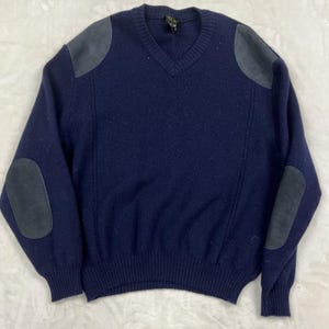 May include: A navy blue V-neck sweater featuring dark grey suede-like patches on the shoulders and elbows. The long-sleeved knit garment has a ribbed neckline, cuffs, and hem, with vertical seam detailing running down the front of the torso.