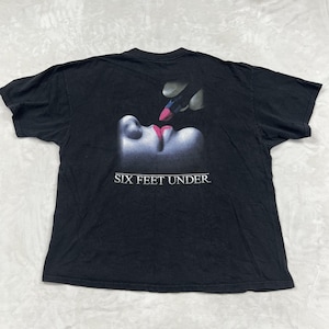 Może przedstawiać: Czarny t-shirt z krótkim rękawem z grafiką przedstawiającą dłoń nakładającą jaskraworóżową szminkę na usta. Pod grafiką znajduje się napis SIX FEET UNDER wydrukowany białymi wielkimi literami. Koszulka posiada okrągły dekolt.