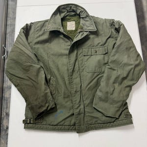 May include: An olive green jacket featuring a pointed collar, a button-up front, and a textured green fleece lining. The jacket includes a single chest pocket with a button flap, long sleeves, and adjustable side tabs at the hem for a fitted look.