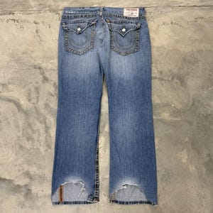 May include: Faded blue denim jeans with a classic five-pocket design. Features decorative stitching on the back pockets and distressed detailing at the hem. The jeans have a straight leg cut and a worn appearance.