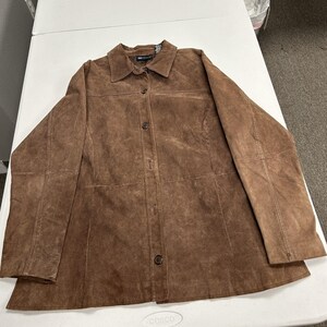 May include: A brown suede button-down shirt jacket featuring a classic collar, long sleeves, and a front button closure. The garment has visible seam detailing across the chest and down the front panels, with a textured finish throughout.