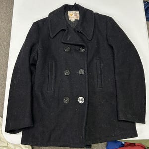 May include: A black double-breasted pea coat featuring a wide collar, long sleeves, and six dark buttons embossed with anchor designs. The interior label reads Fox Knapp Sportswear. The coat has two vertical slit pockets on the front panel.